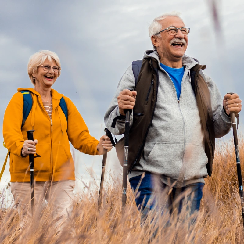 Active senior couple hiking through golden field with walking poles, wearing outdoor gear and smiling while enjoying nature together